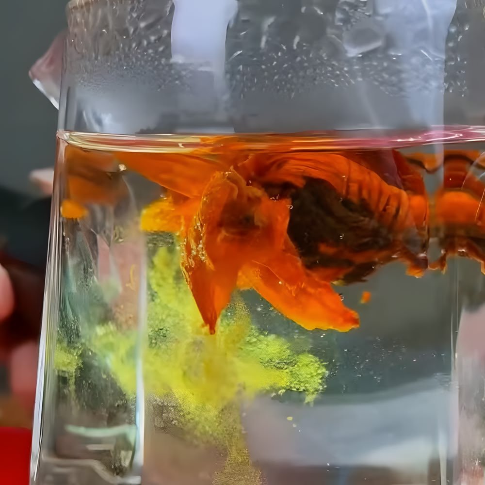 A close-up shot of a Lily Pagoda Black Blooming Tea steeping in a clear glass vessel. The orange lily flower has unfurled, and a visible cloud of natural golden pollen is actively scattering and dissolving into the water beneath the dark Dianhong tea leaves.
