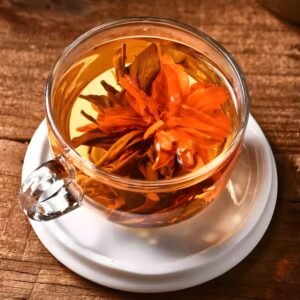 Close-up view of a clear glass cup holding a fully unfurled Lily Pagoda Bloom. Vibrant orange lily petals reveal a heart of dark Dianhong black tea leaves. The tea liquor is a rich, warm amber color, set on a rustic wooden table.