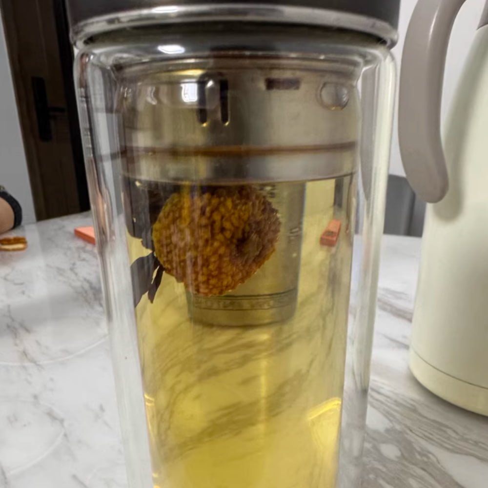 A lifestyle photo showing Chestnut Rose tea brewing in a tall double-walled glass tumbler on a marble table next to a kettle.