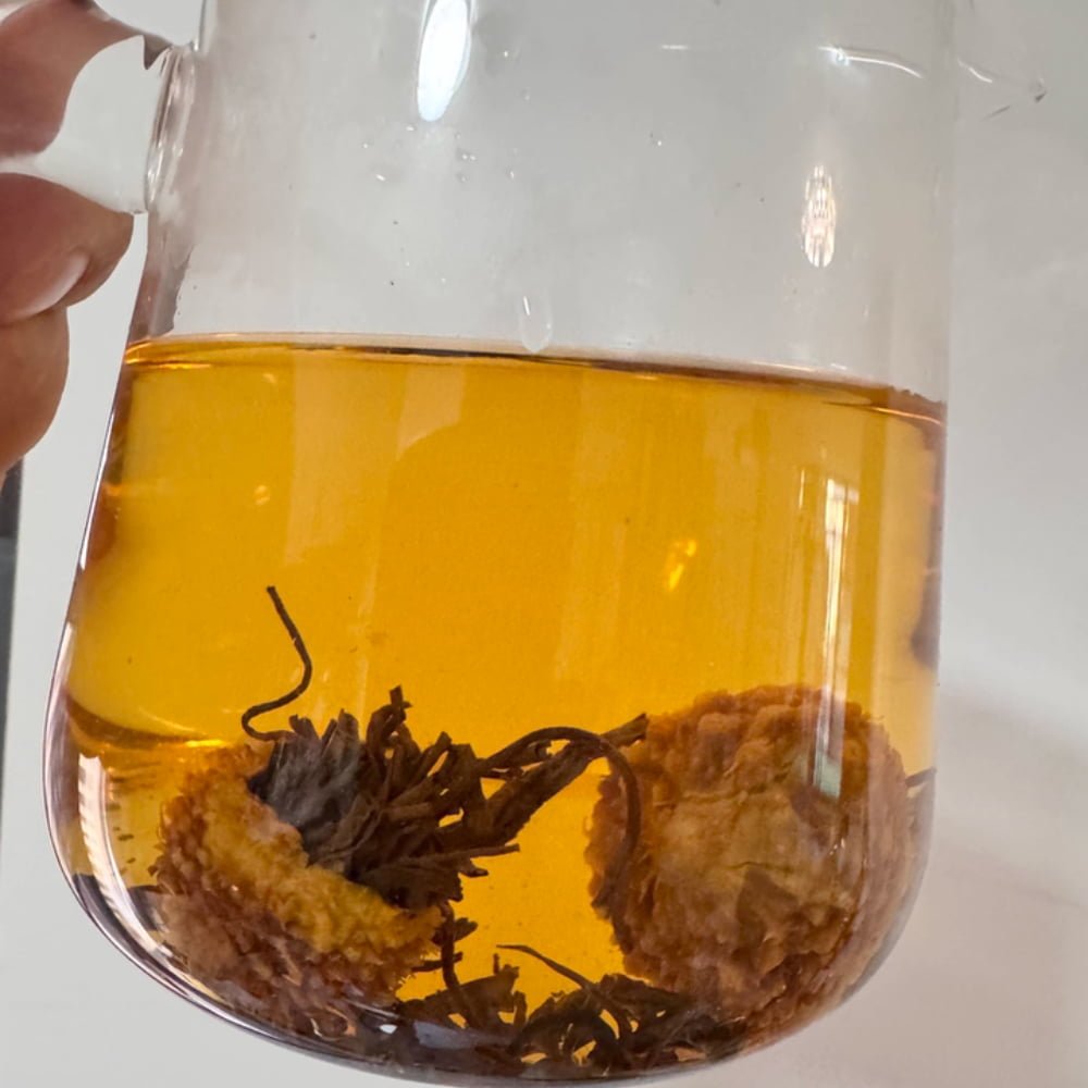 A customer review photo showing a hand holding a glass pitcher of brewed Chestnut Rose tea. The liquid is a clear amber color, with two whole fruits and expanded tea leaves visible at the bottom.