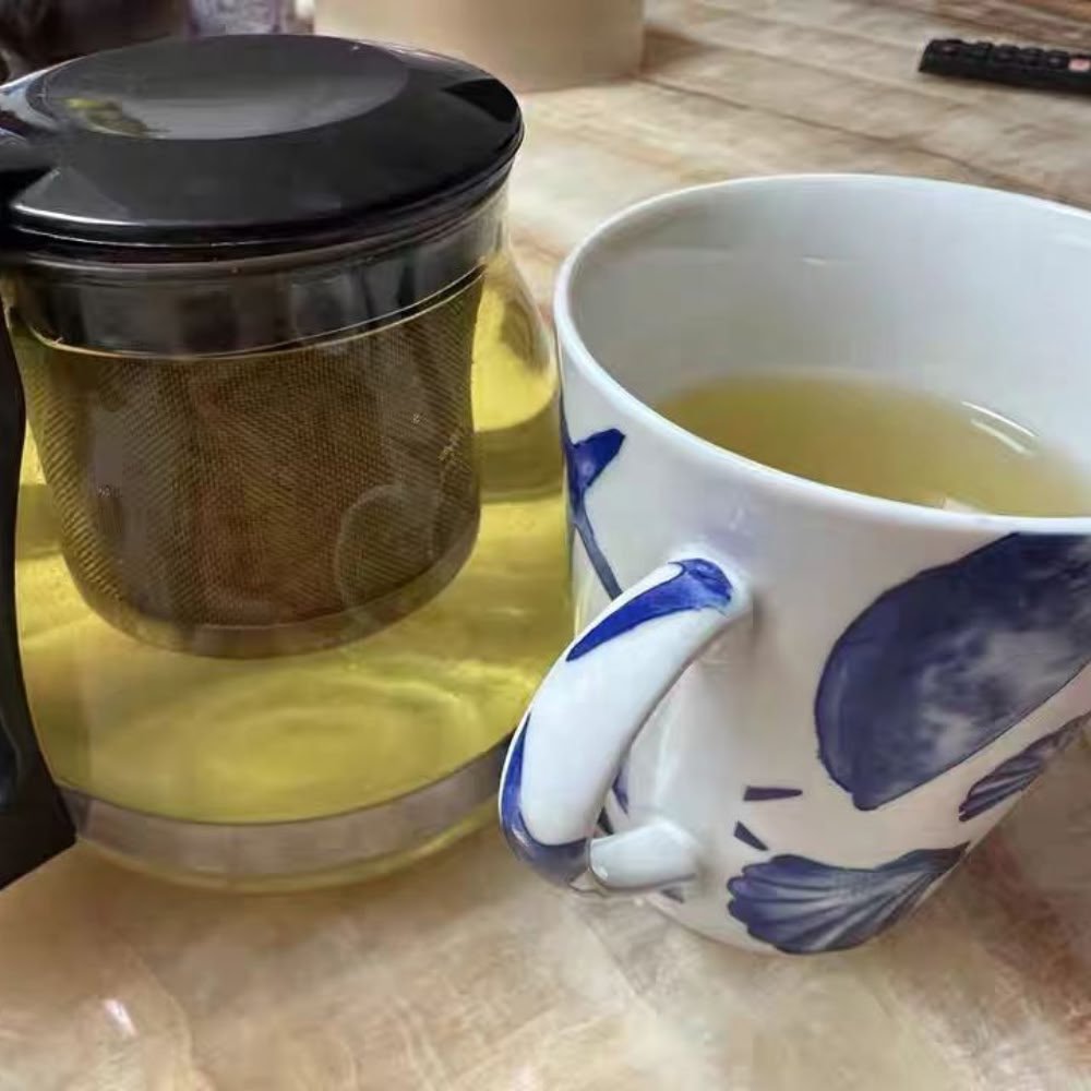 A glass teapot with an infuser next to a blue and white ceramic mug filled with golden Nutty Tartary Buckwheat tea on a table.