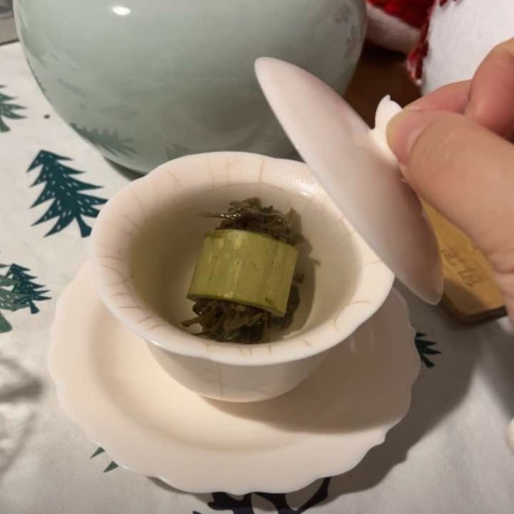 A hand lifting the lid of a traditional white porcelain gaiwan, revealing raw Pu-erh tea housed inside a natural bamboo section, ready to brew a bright golden infusion.