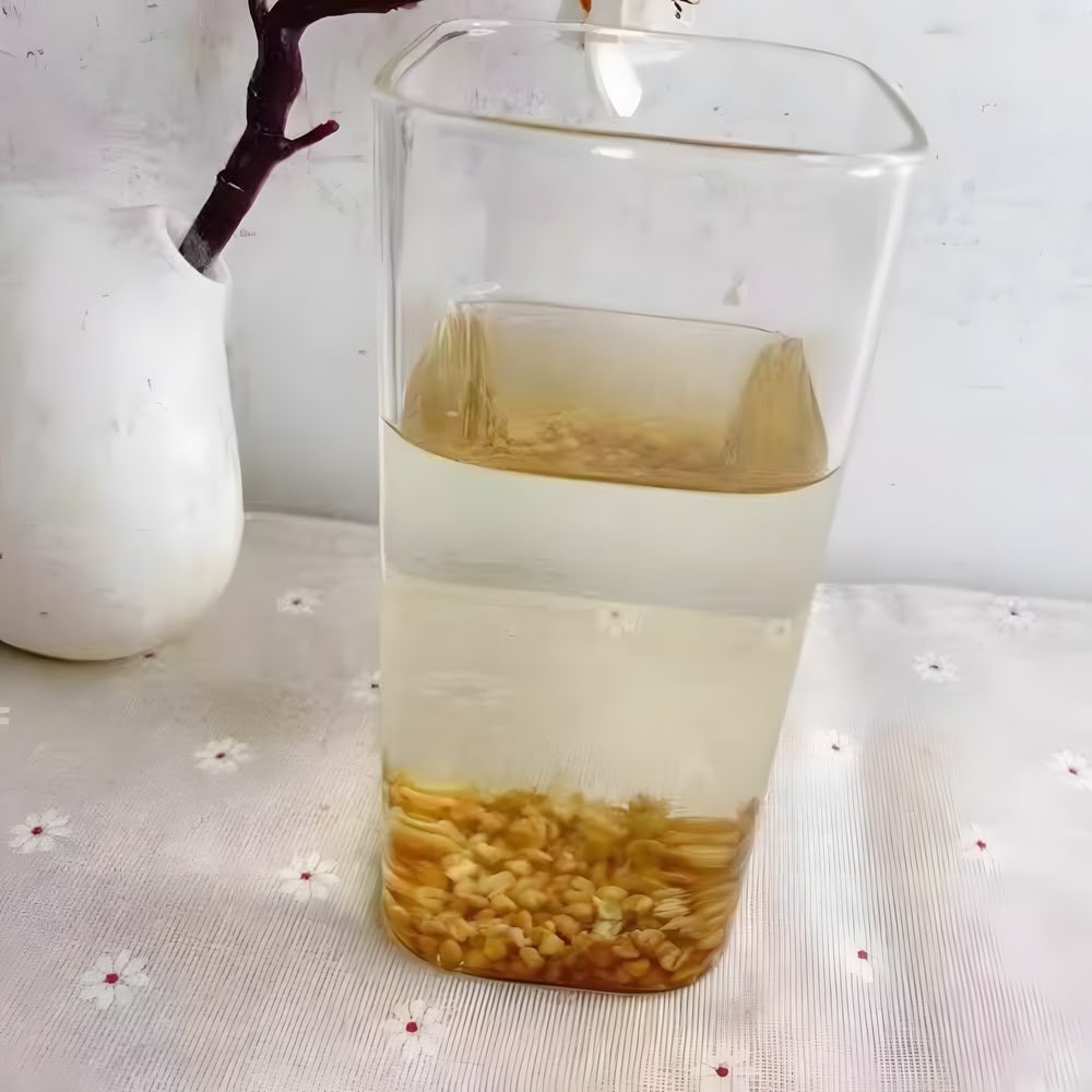 A tall, clear glass pitcher filled with steeped Nutty Tartary Buckwheat tea and soft kernels resting at the bottom, placed on a floral tablecloth.