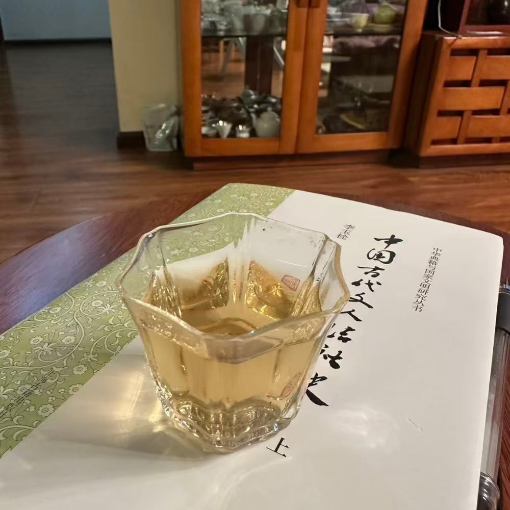 A cozy evening customer photo showing a beautiful angular glass of Dongzisu tea resting on a Chinese book, with warm home decor in the background.