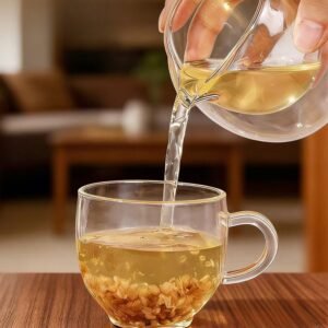 A sunlit aesthetic close-up shot of a hand pouring clear, golden Nutty Tartary Buckwheat tea from a glass pitcher into a matching cup filled with steeped kernels.
