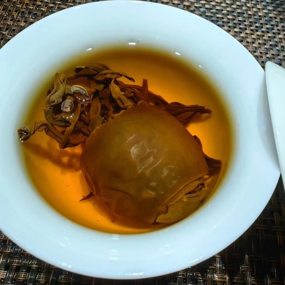 A top-down view of a white porcelain teacup containing golden tea liquor. The steeped tree tomato shell and fully unfurled, whole tea leaves rest beautifully in the clear infusion.
